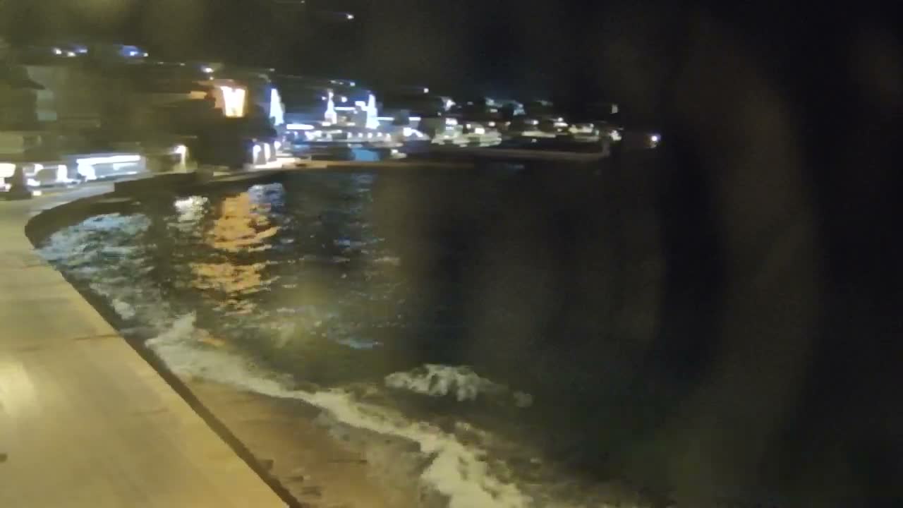 A concrete pier lined with parked cars extends into a vast expanse of turquoise water under a sky marked by a mix of bright sunshine, white fluffy clouds, and darker, heavier storm clouds, with distant land visible on the horizon.