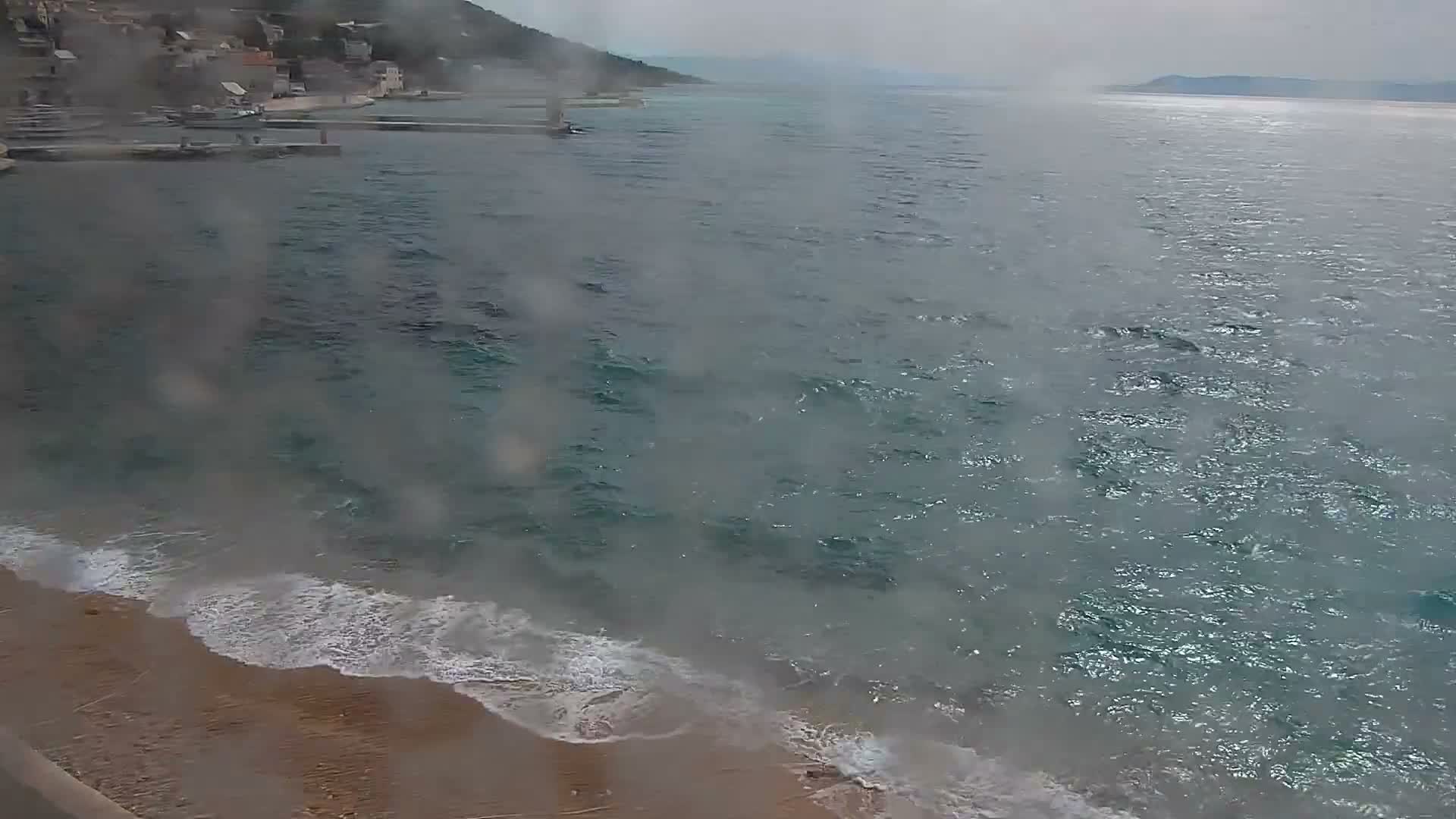 A concrete pier lined with parked cars extends into a vast expanse of turquoise water under a sky marked by a mix of bright sunshine, white fluffy clouds, and darker, heavier storm clouds, with distant land visible on the horizon.
