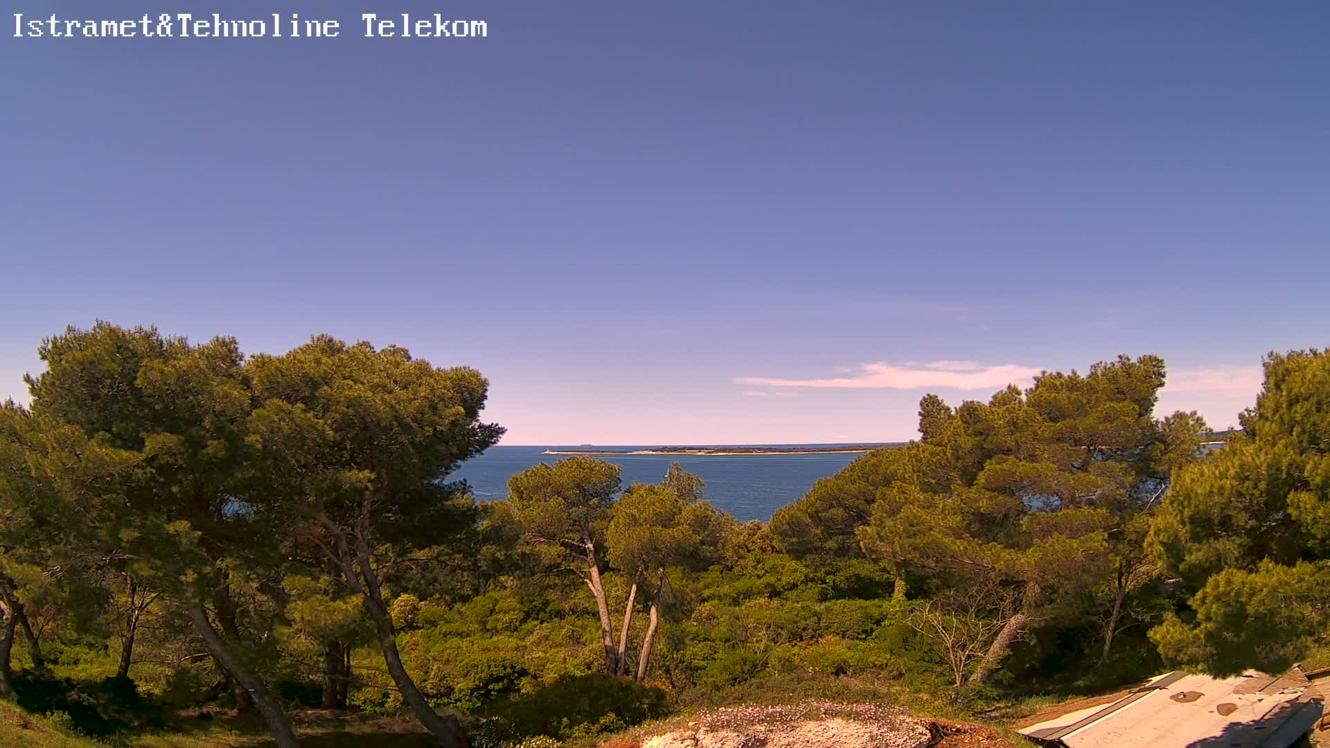 A clear, sunny day reveals a tranquil coastal scene of lush green vegetation, including pine trees, overlooking a calm blue sea with a distant landmass visible on the horizon.