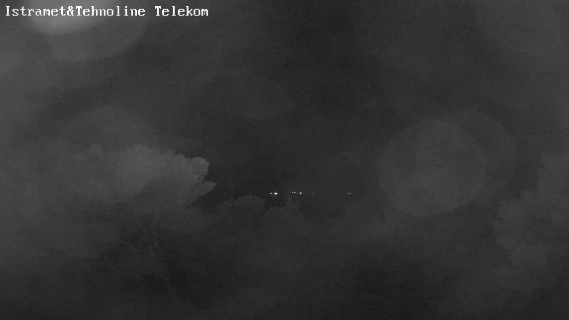 A nighttime, cloudy view shows a few distant lights beyond a dark, overcast sky.
