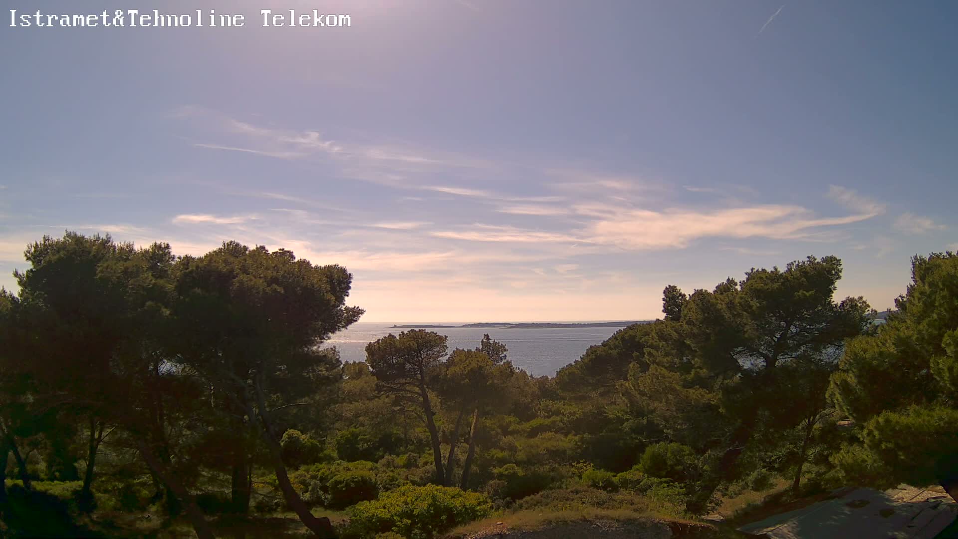 A calm sea is visible beyond a hillside covered in dark green pine trees under a mostly clear sky with a few wispy clouds.