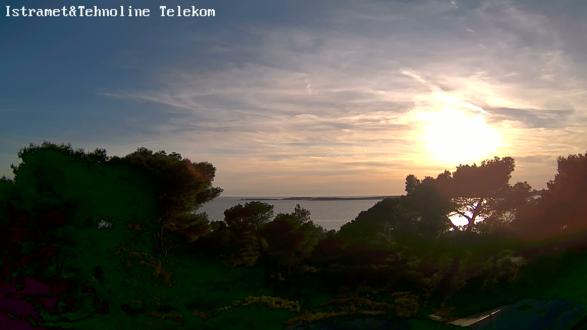 A partly cloudy sunset over a calm body of water, viewed from a hilltop covered in dark green trees.