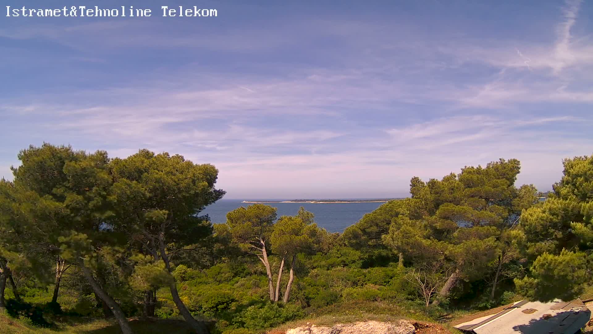 A partly cloudy blue sky overlooks a calm sea and a lush green landscape of pine trees and low shrubs.