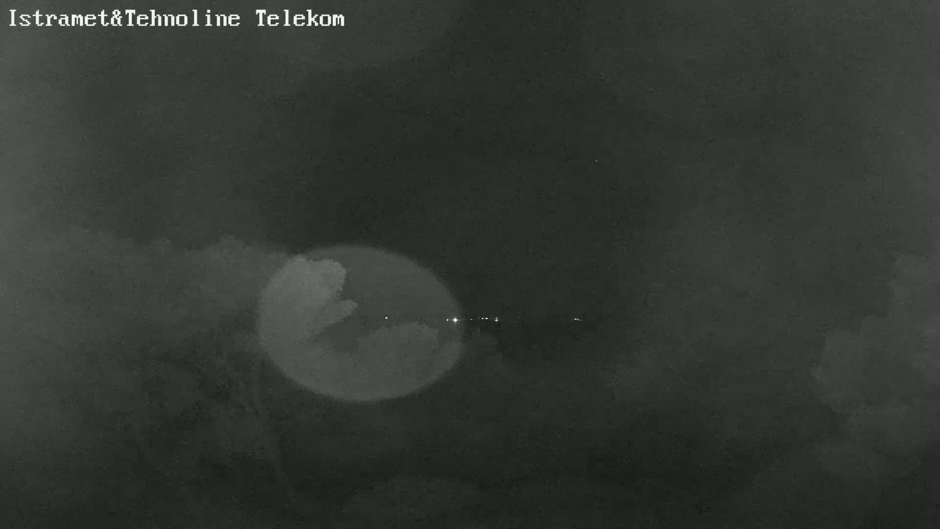 A nighttime, blurry image shows a dark landscape with a few distant lights and a bright, hazy circle in the foreground.