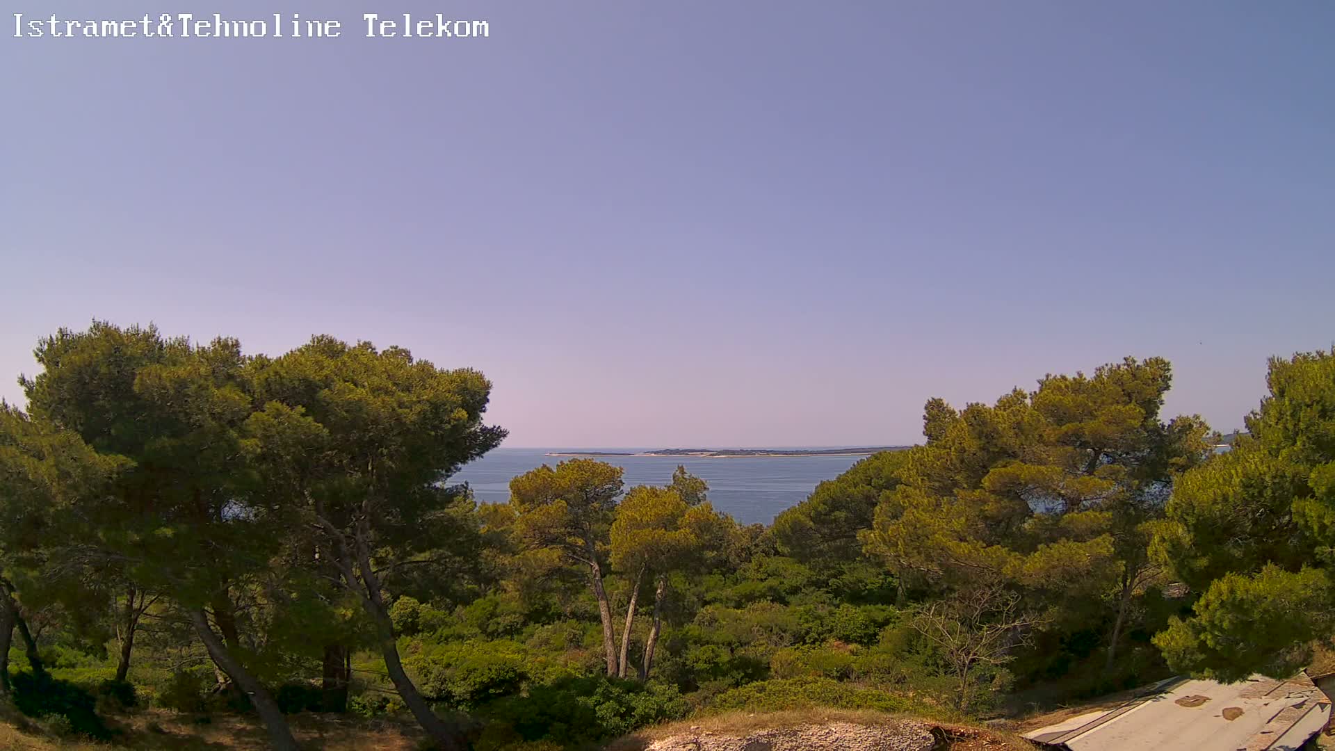 A clear, sunny day reveals a tranquil coastal scene with lush green vegetation and a calm blue sea, featuring a small island in the distance.