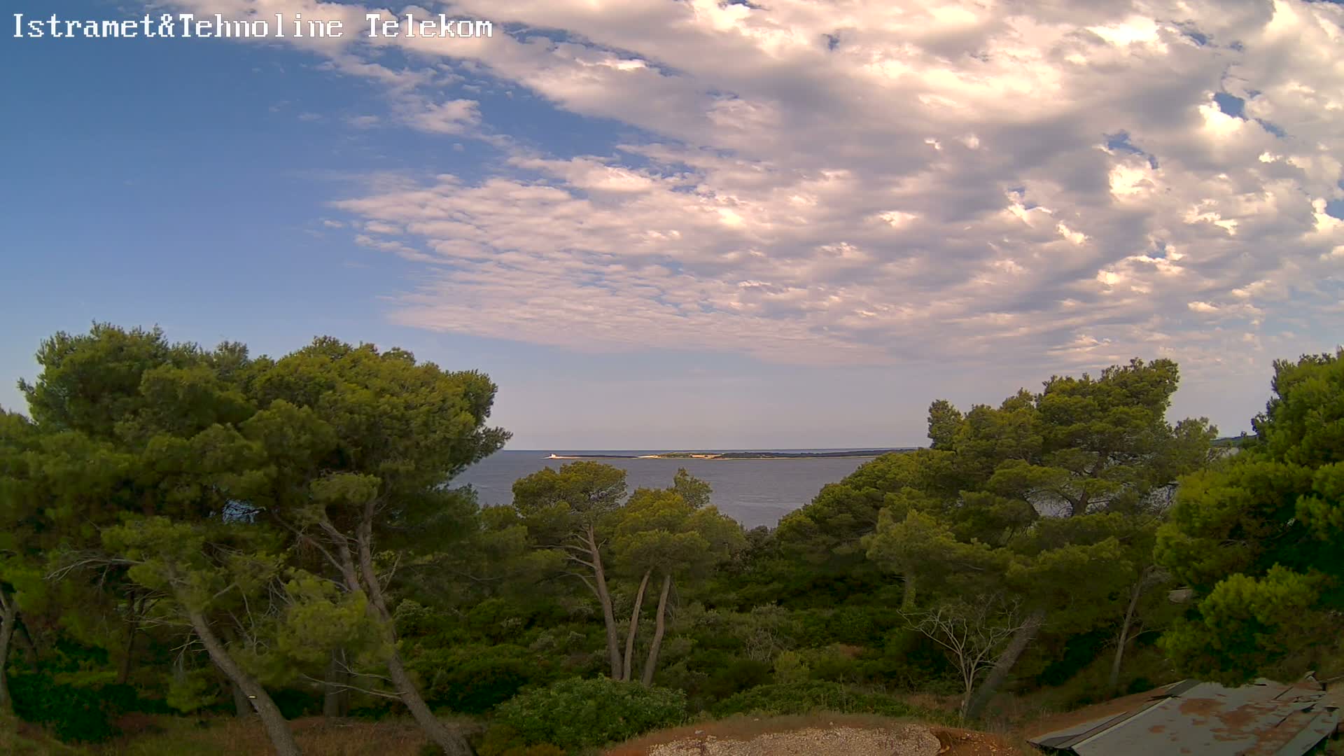 A partly cloudy sky overlooks a calm sea and a lush green landscape of pine trees.
