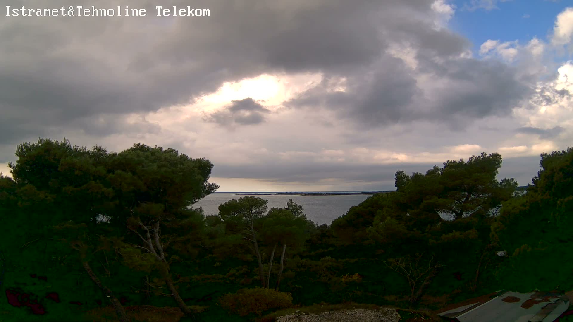 Dense evergreen trees frame a calm body of water and distant land under a dramatic, partly cloudy sky with heavy clouds and bright patches of sunlight breaking through.