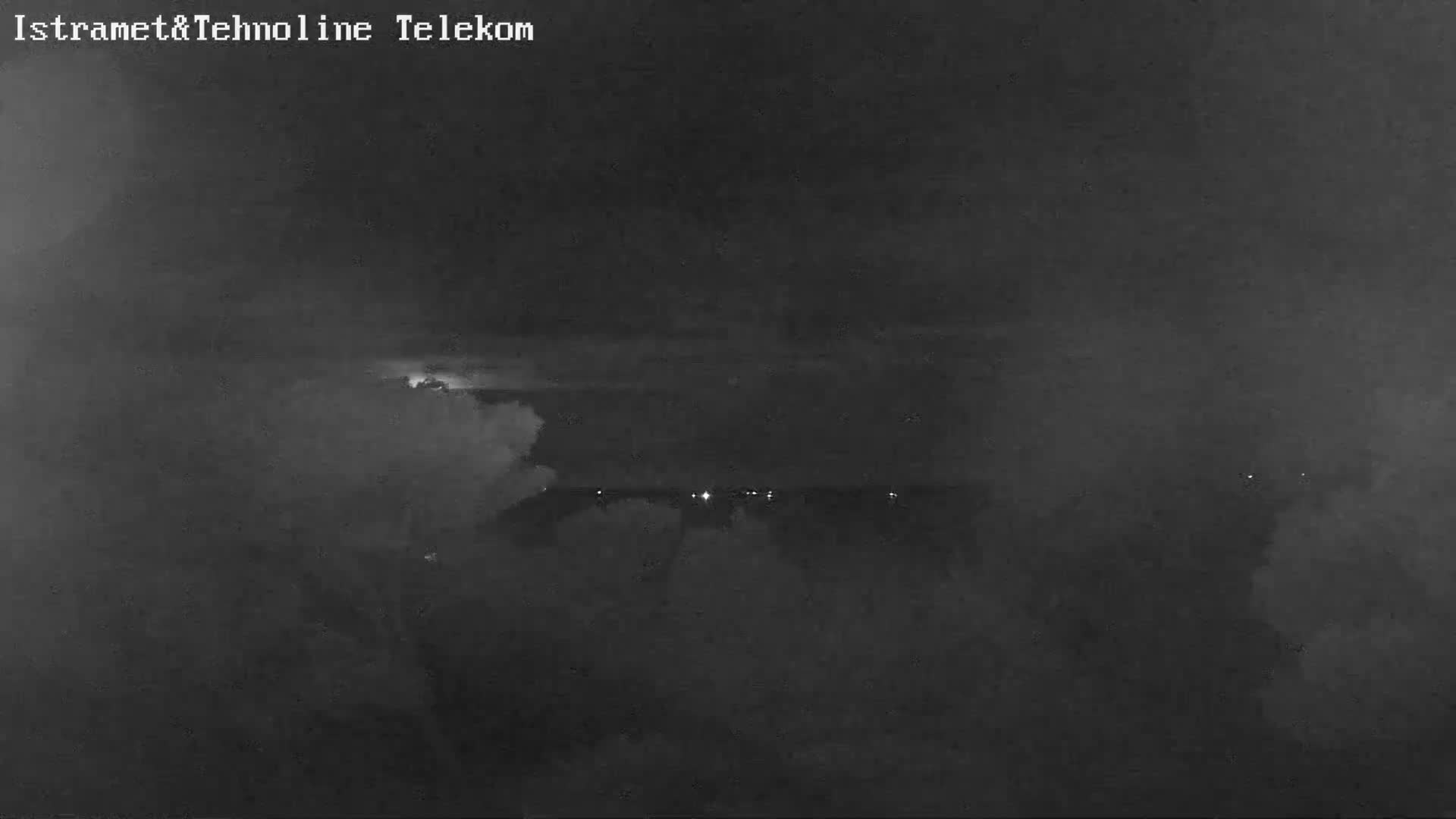 A dark, stormy night scene displays a cloudy sky illuminated by flashes of lightning, with a faint line of distant lights visible below.