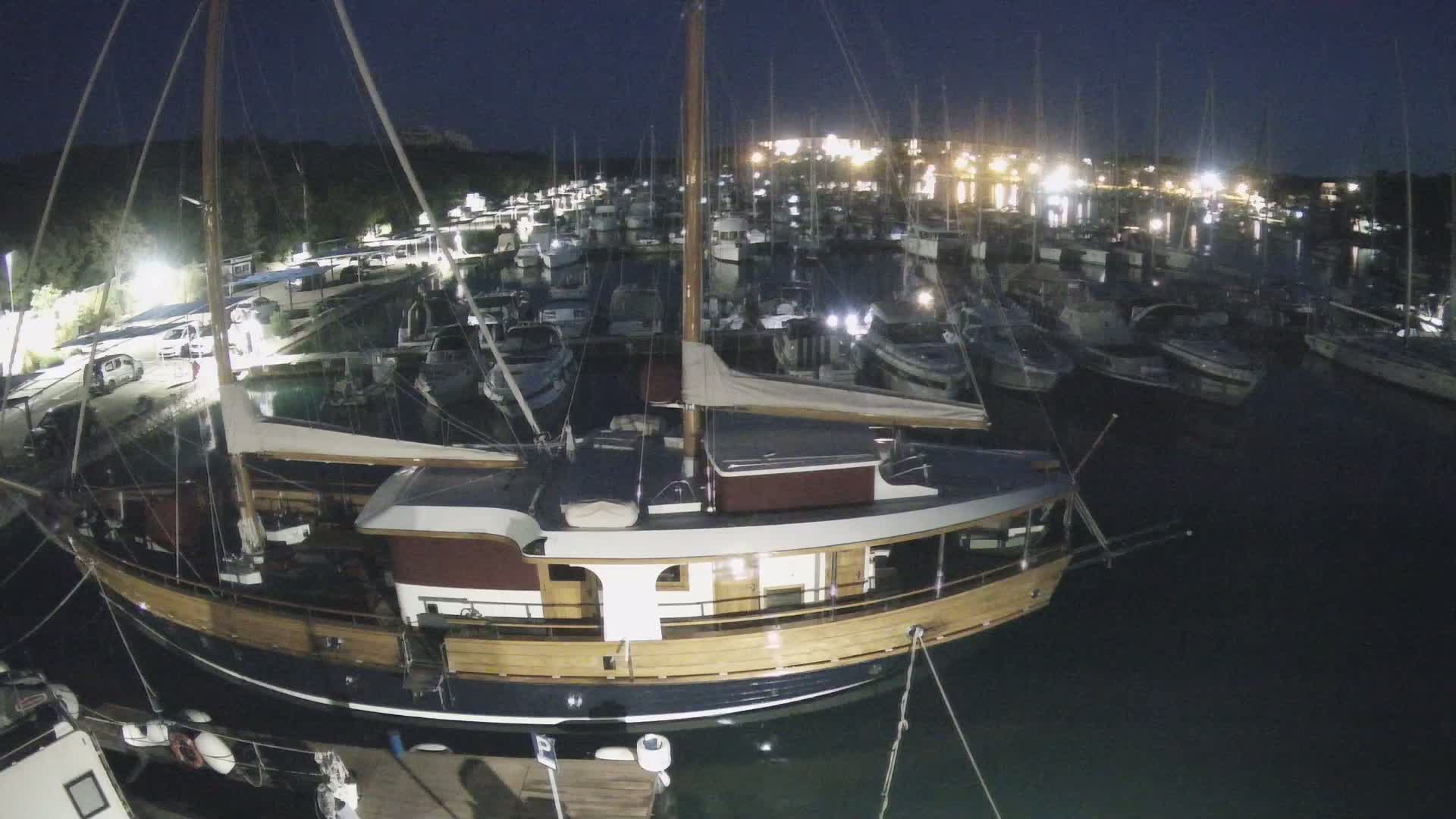 A large wooden sailboat is moored in a marina at night, surrounded by many other boats under a dark sky.