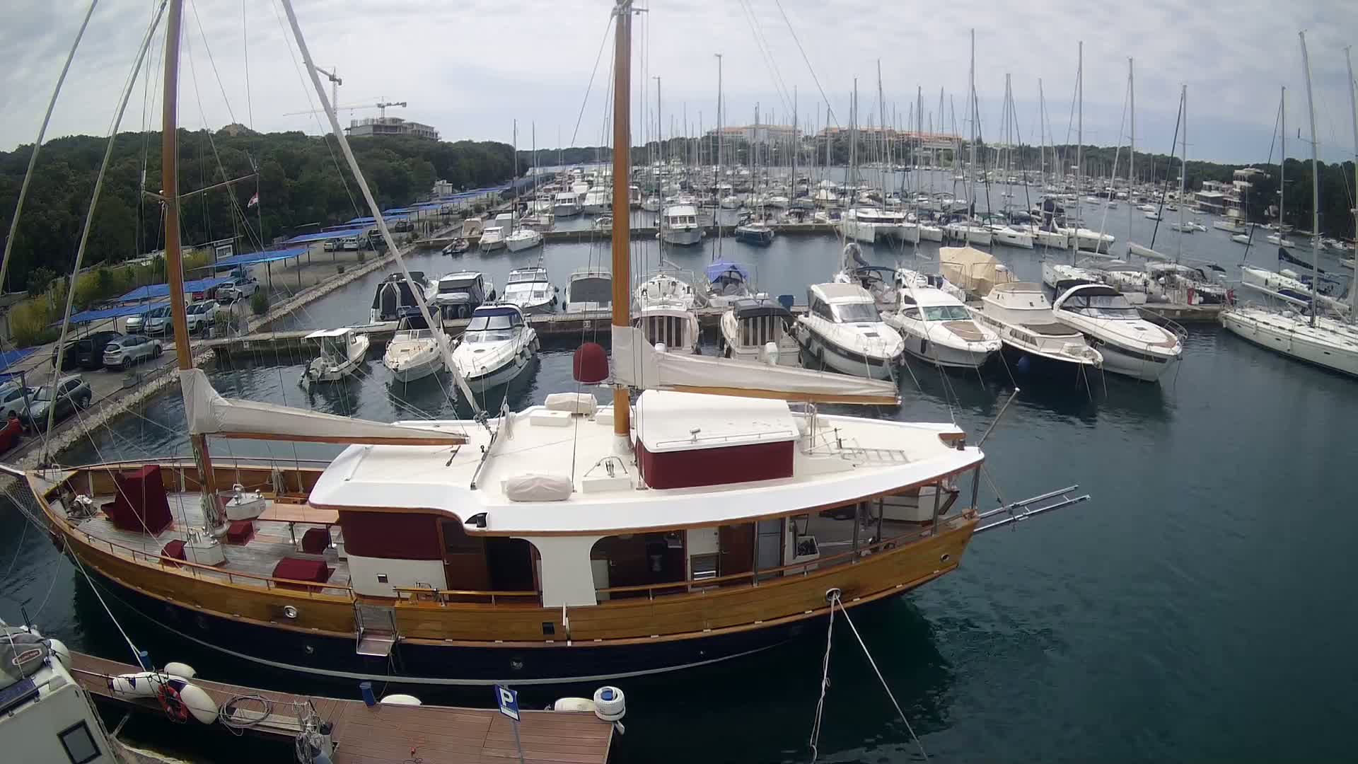 A large wooden sailboat is moored in a marina filled with numerous other sailboats and motorboats on an overcast day.