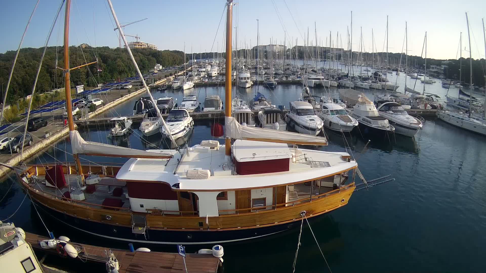 A large wooden sailboat is docked in a marina filled with numerous other sailboats and motorboats on a calm, sunny day.