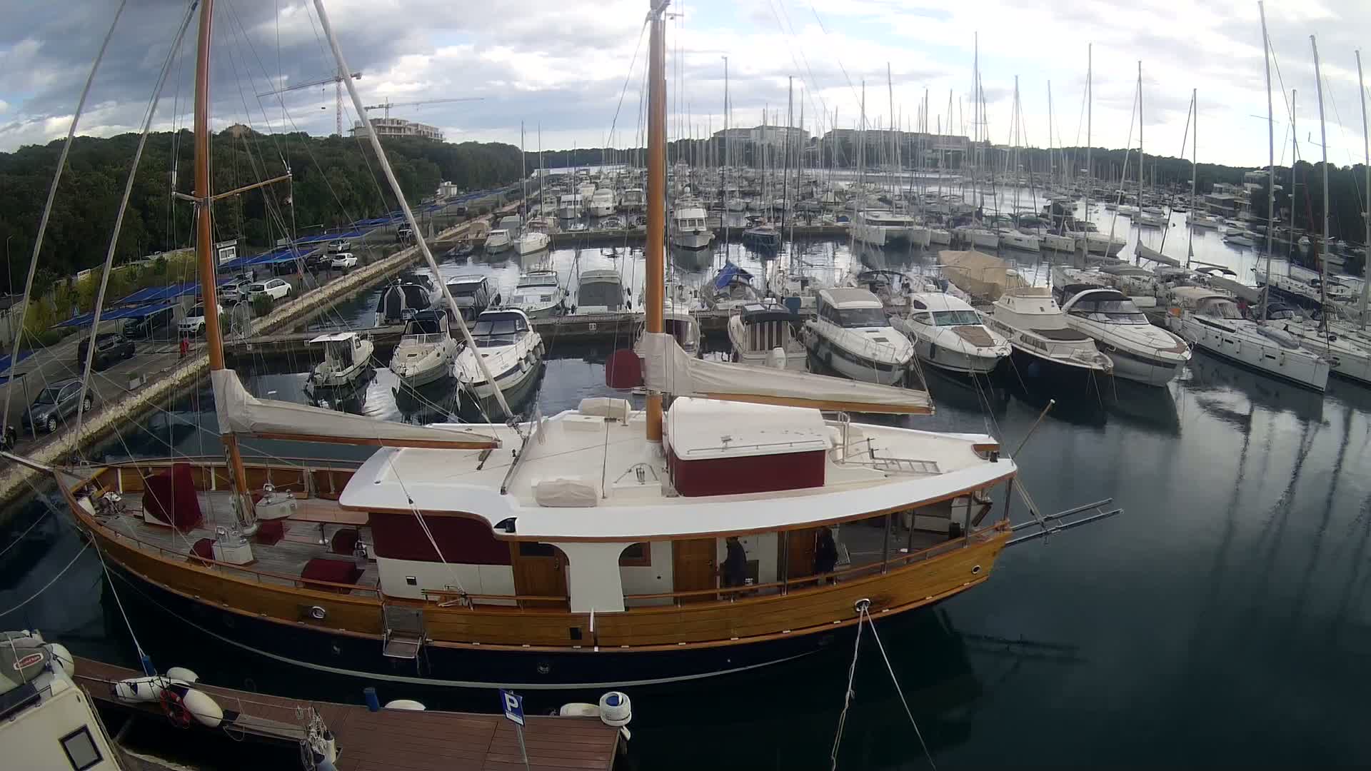 A large wooden sailboat is docked in a marina filled with many other boats on an overcast day.