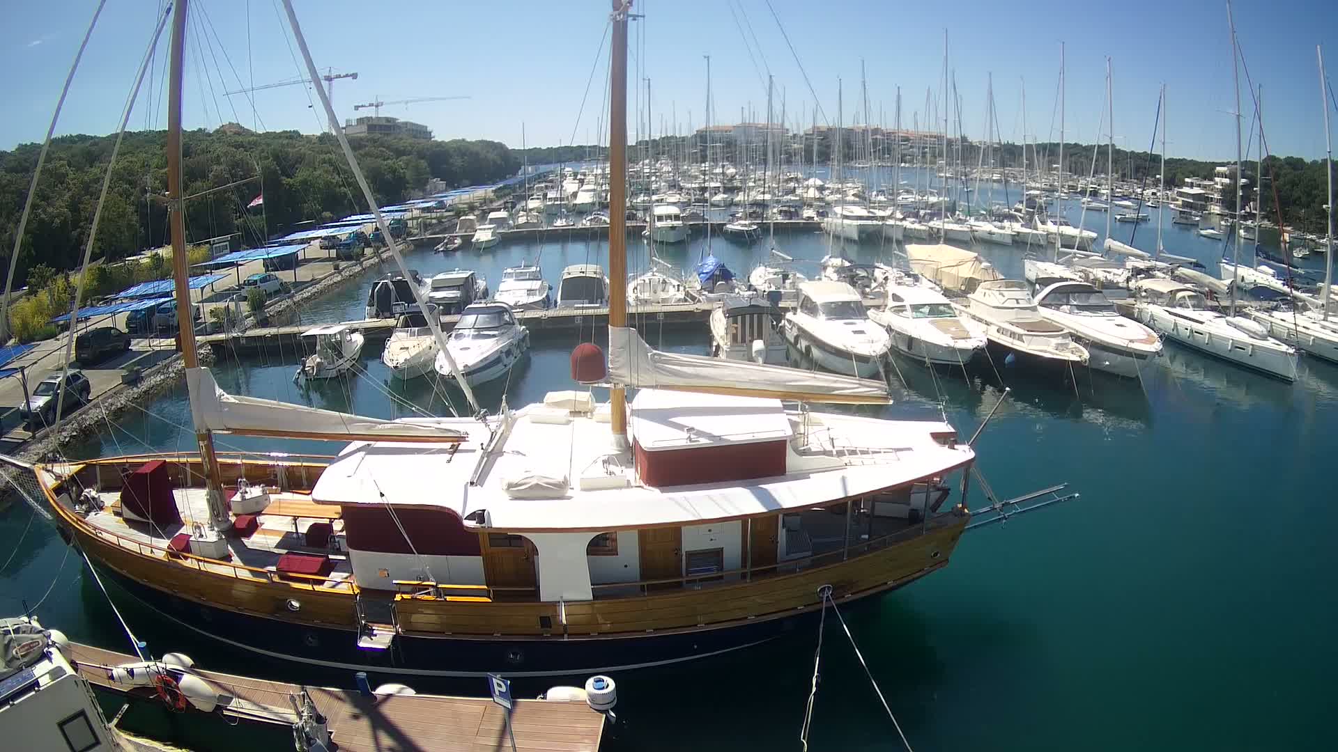 A large wooden sailboat is docked in a marina filled with numerous other boats on a sunny day.