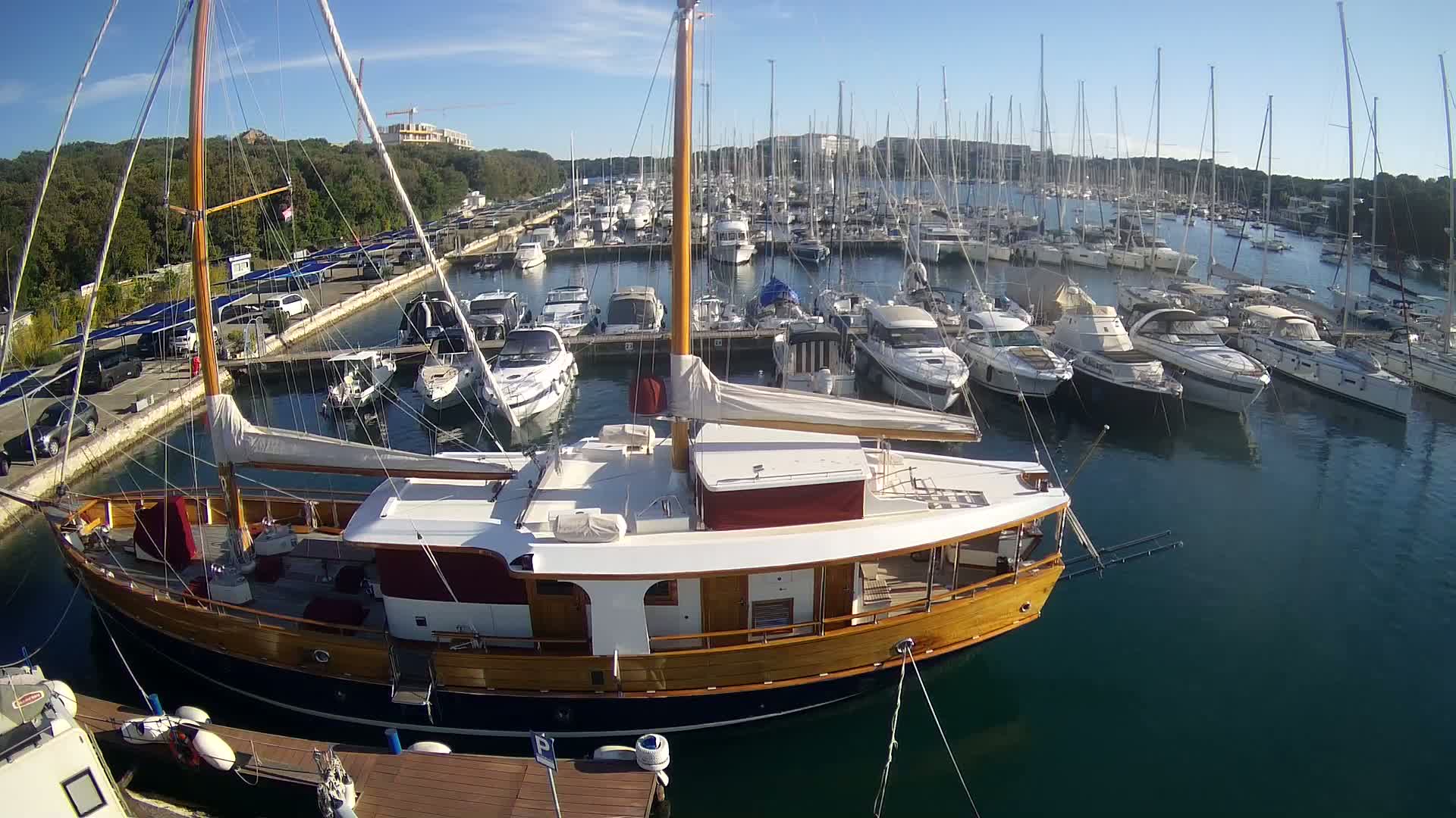 A large wooden sailboat is docked in a marina filled with numerous other boats on a sunny day.