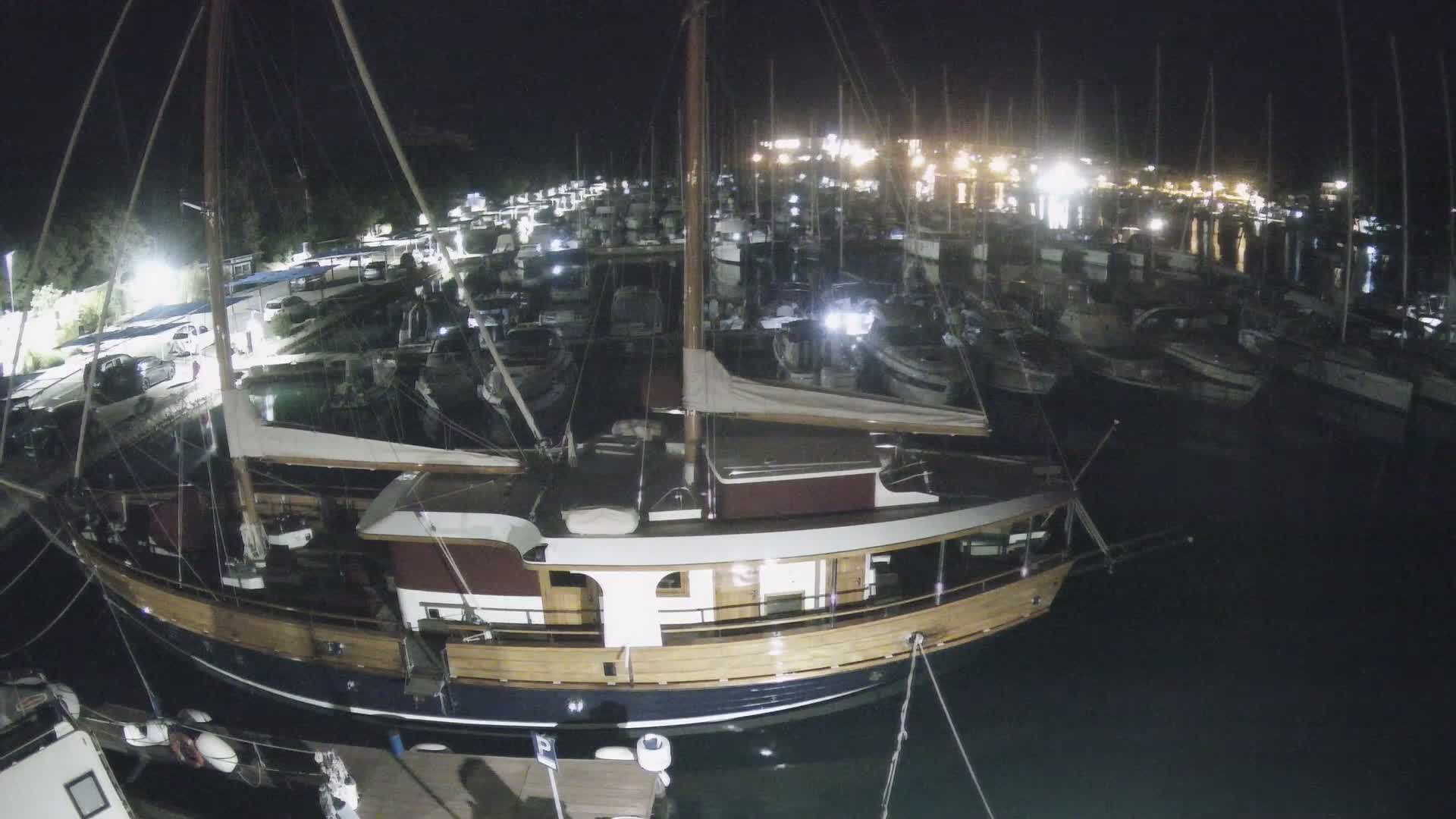 A large wooden sailboat is moored at a marina at night, surrounded by many other boats and illuminated by lights on the shore.