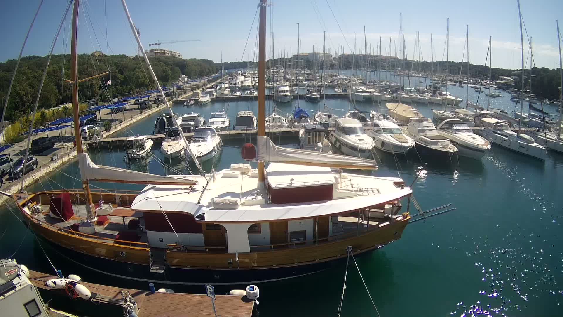 A large wooden sailboat is docked in a marina filled with numerous other boats on a sunny day.