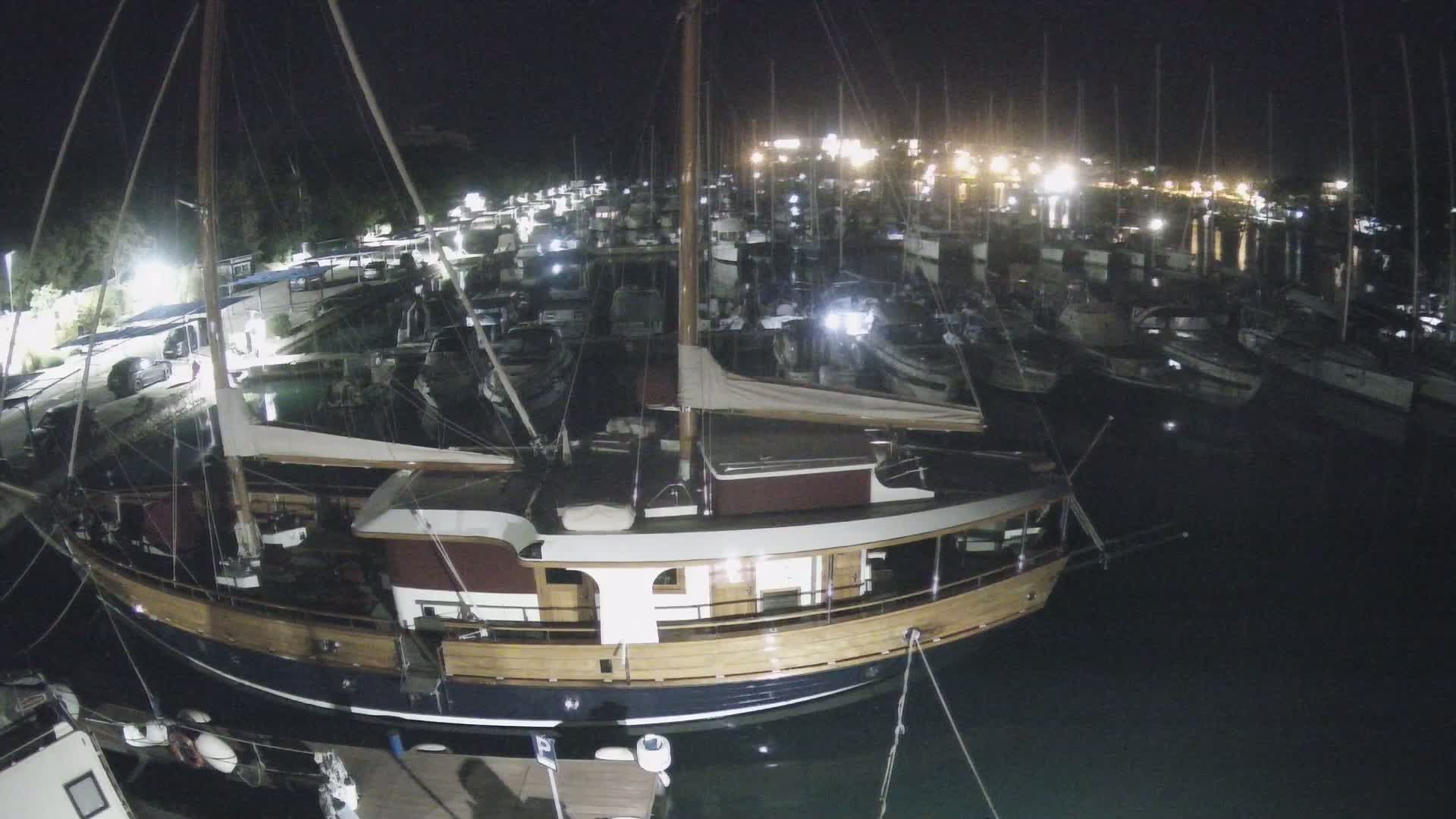 A large wooden boat is moored in a marina at night, surrounded by numerous other boats under a dark sky.