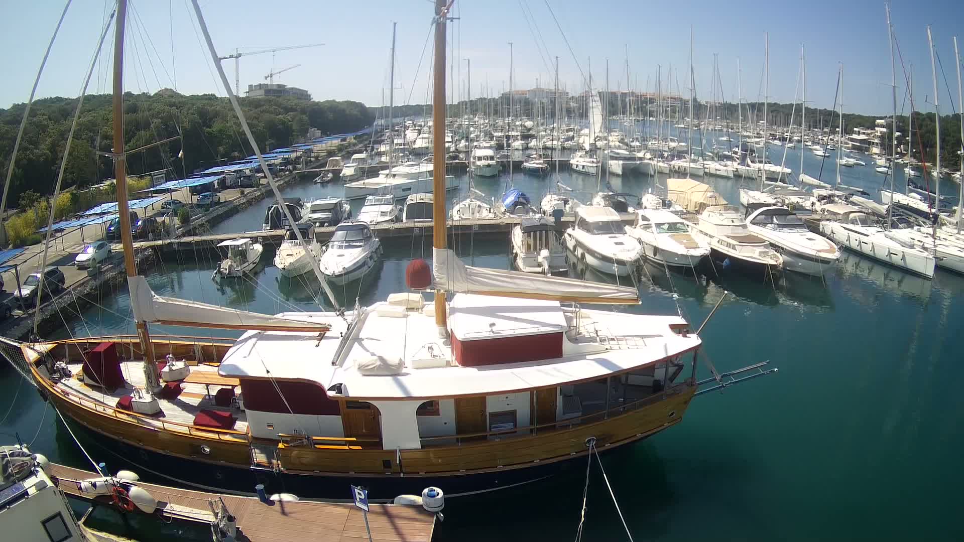 A large wooden sailboat is docked in a marina filled with numerous other sailboats and motorboats on a sunny day.