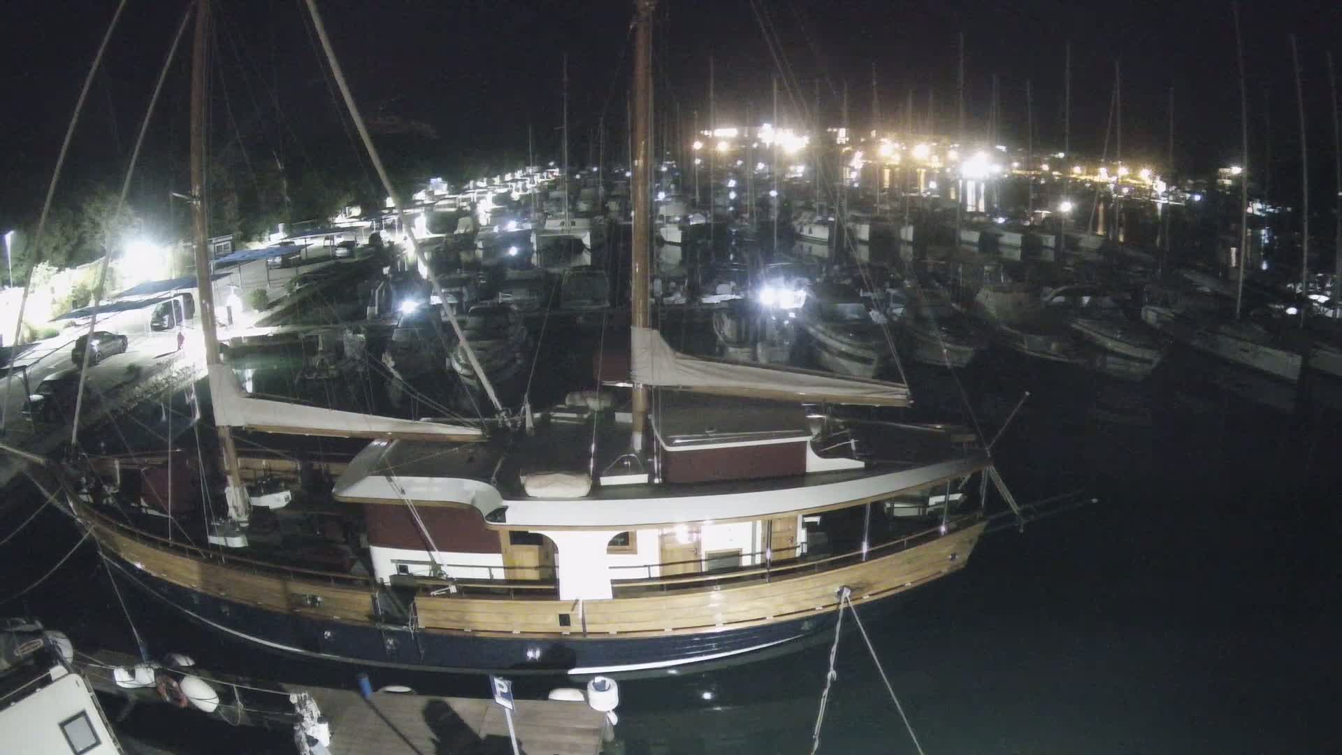 A large wooden sailboat is docked at night in a marina filled with many other boats, all illuminated by artificial lights.
