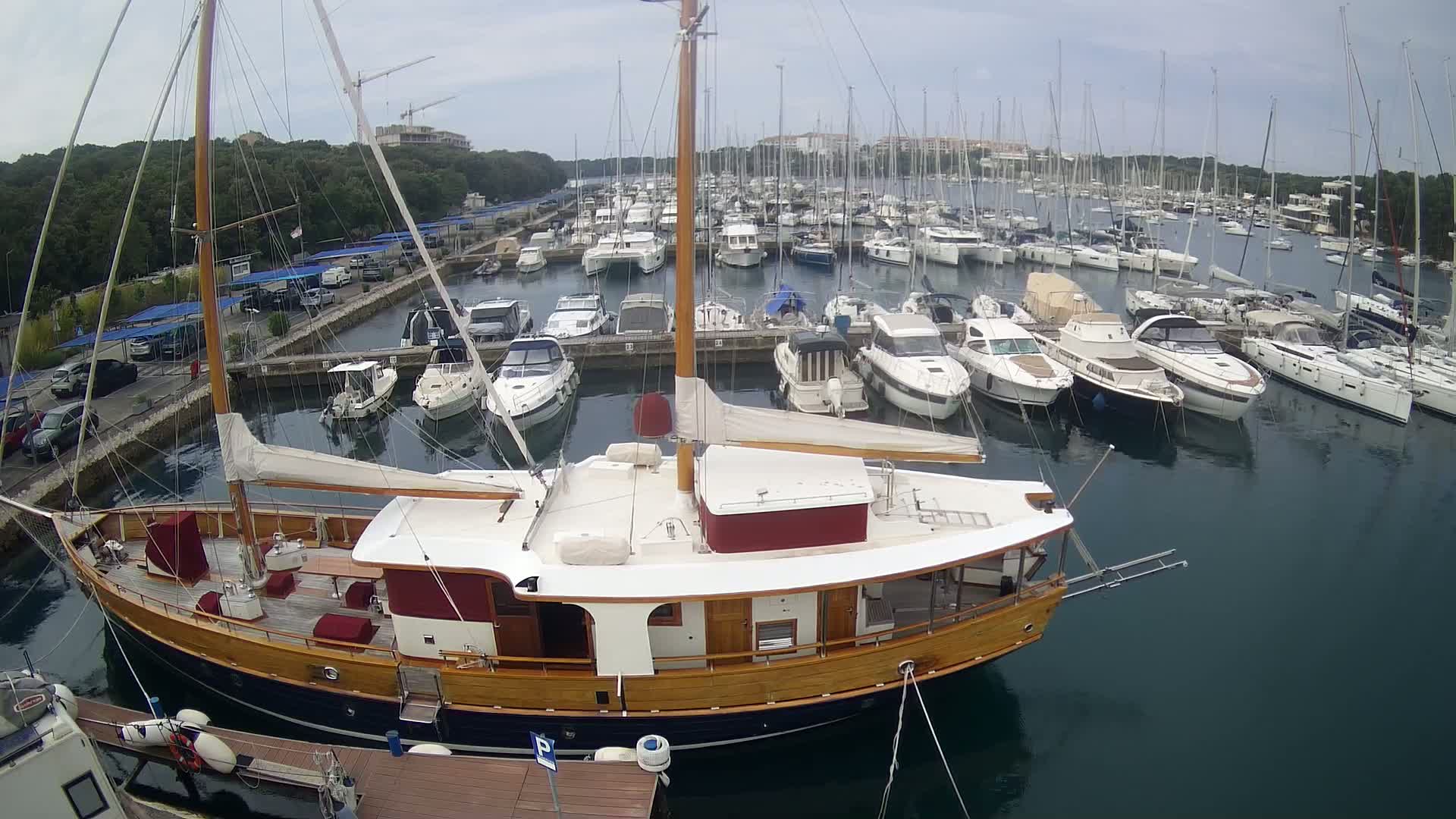 A large wooden sailboat is docked in a marina filled with numerous other boats on an overcast day.