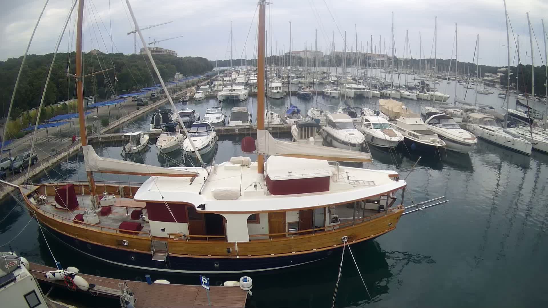 A large wooden sailboat is docked in a marina filled with numerous other boats on an overcast day.