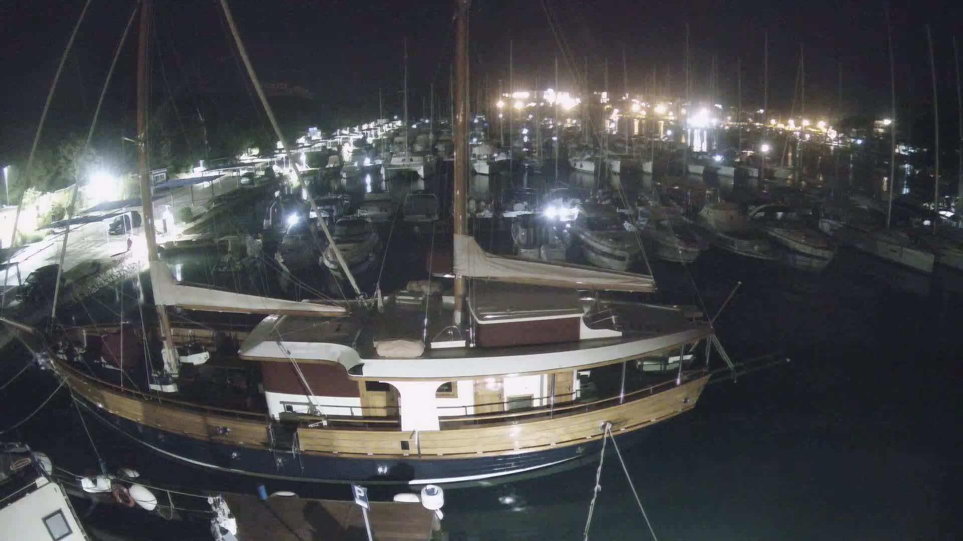 A large wooden sailboat is moored in a marina at night, surrounded by numerous other boats under dark, clear skies.