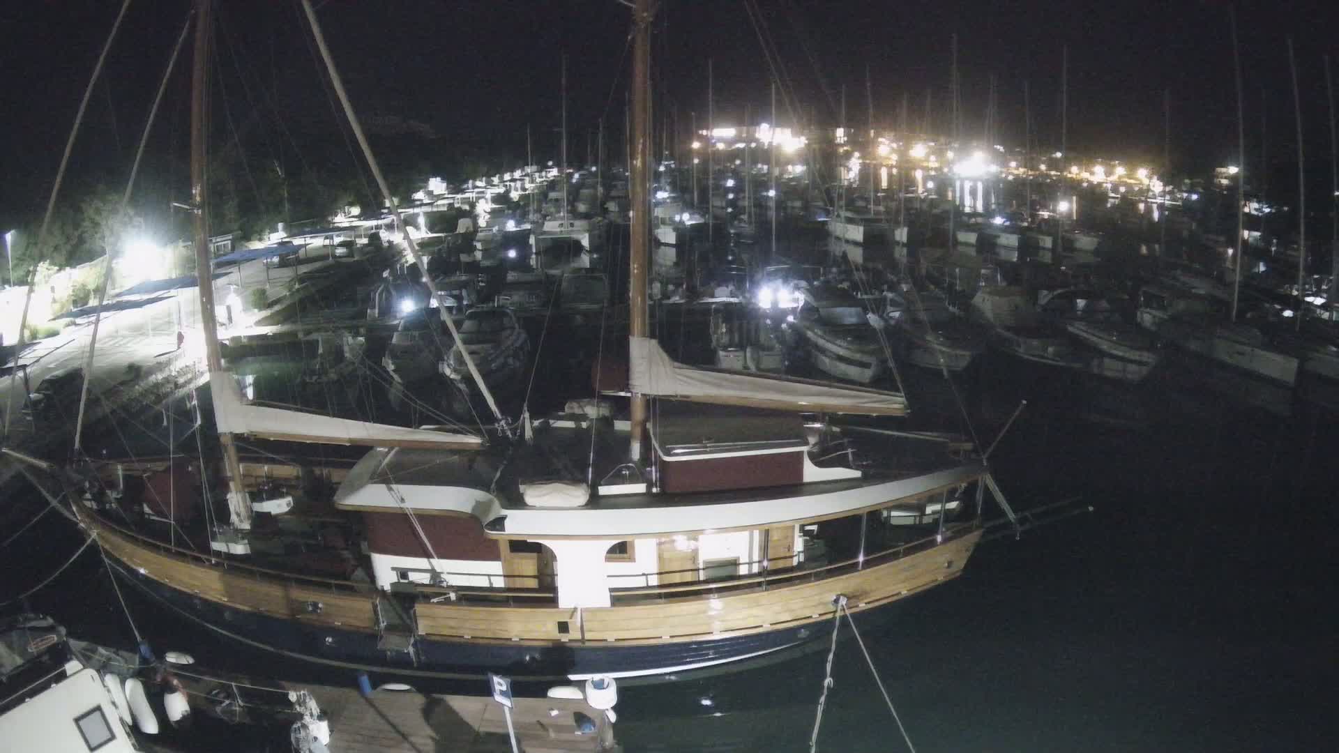 A large wooden sailboat is docked at night among many other smaller boats in a marina illuminated by artificial lights.