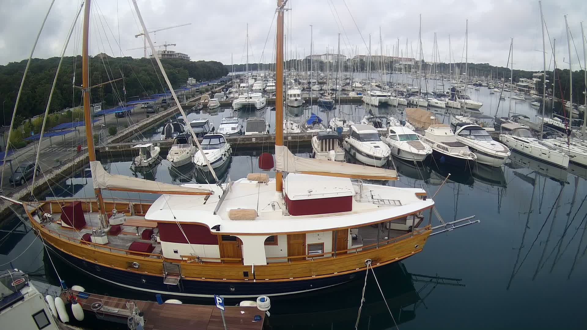 A wooden sailboat is docked in a marina filled with numerous other sailboats and motorboats under an overcast sky.