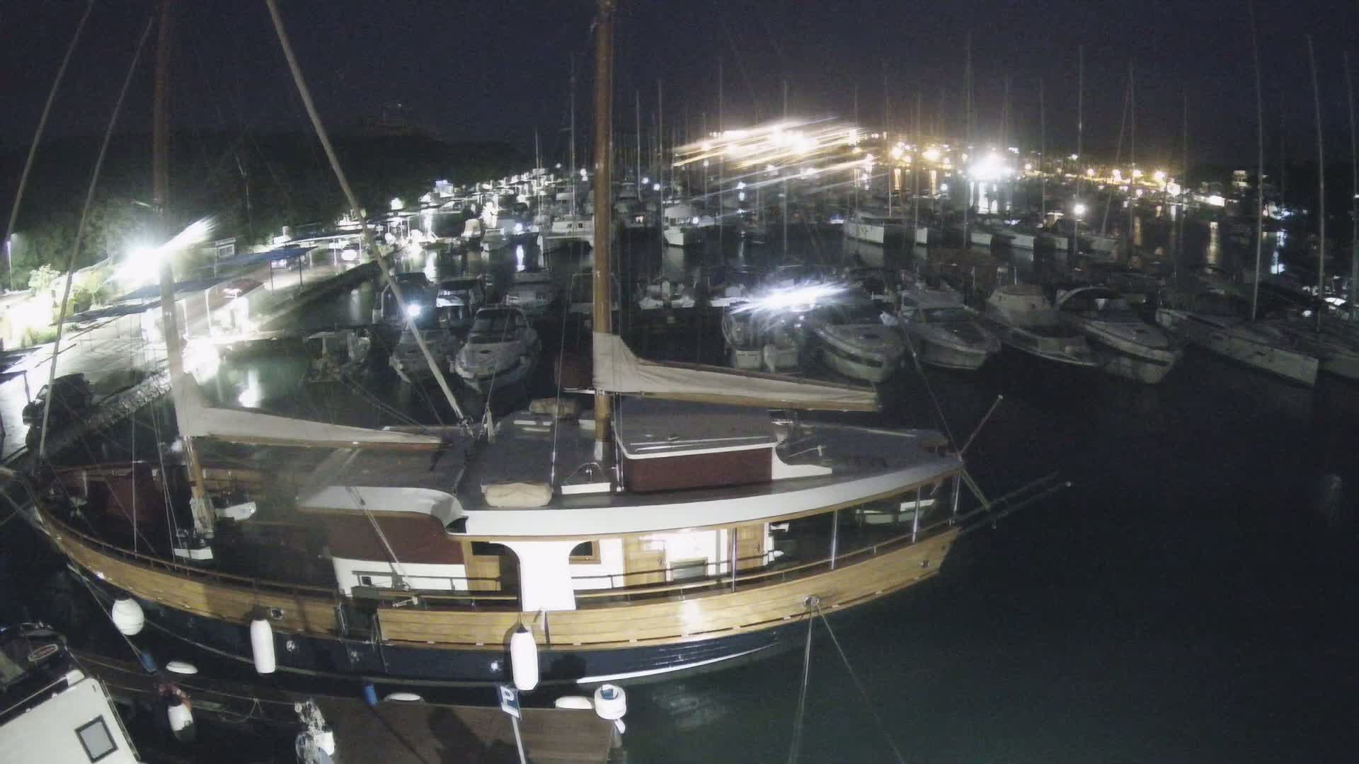 A large wooden sailboat is moored in a marina at night, surrounded by many other boats under dark, calm conditions.