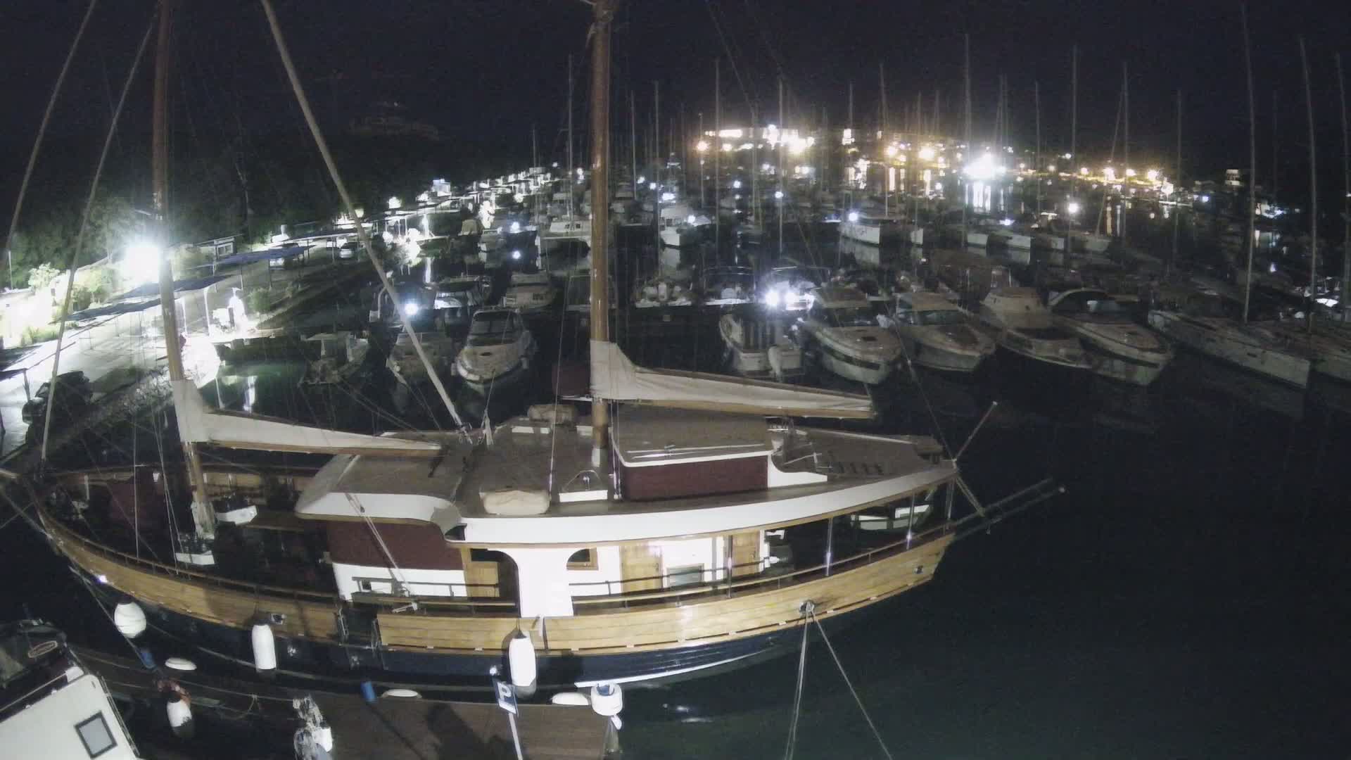 A large wooden boat is moored at a marina at night, surrounded by many smaller boats under a dark sky.