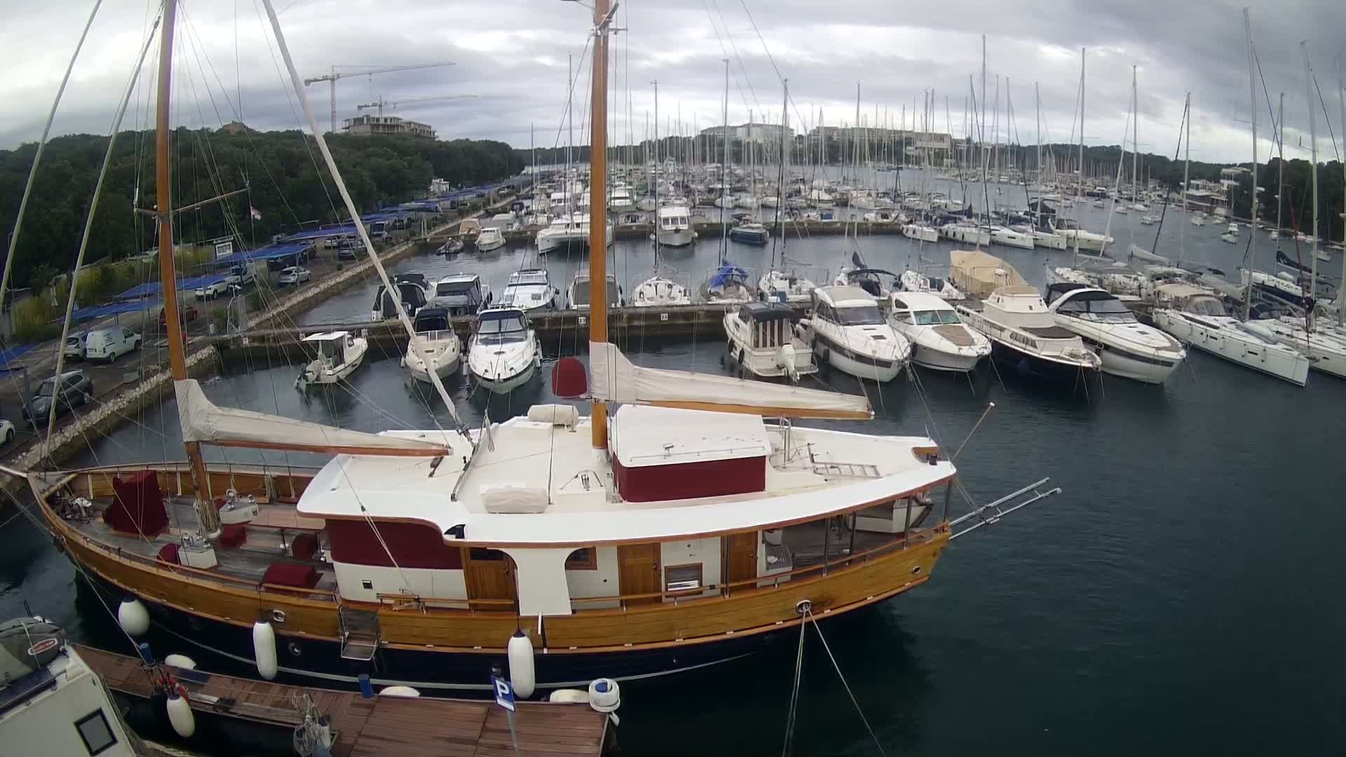 A large wooden sailboat is docked in a marina filled with numerous other boats on an overcast day.
