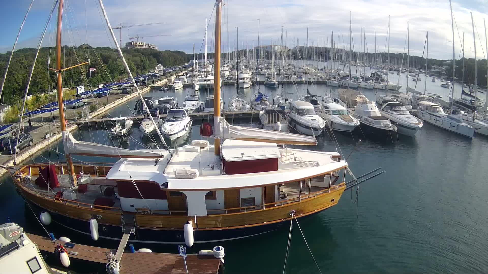 A large wooden sailboat is docked in a marina filled with numerous other boats on a sunny day.