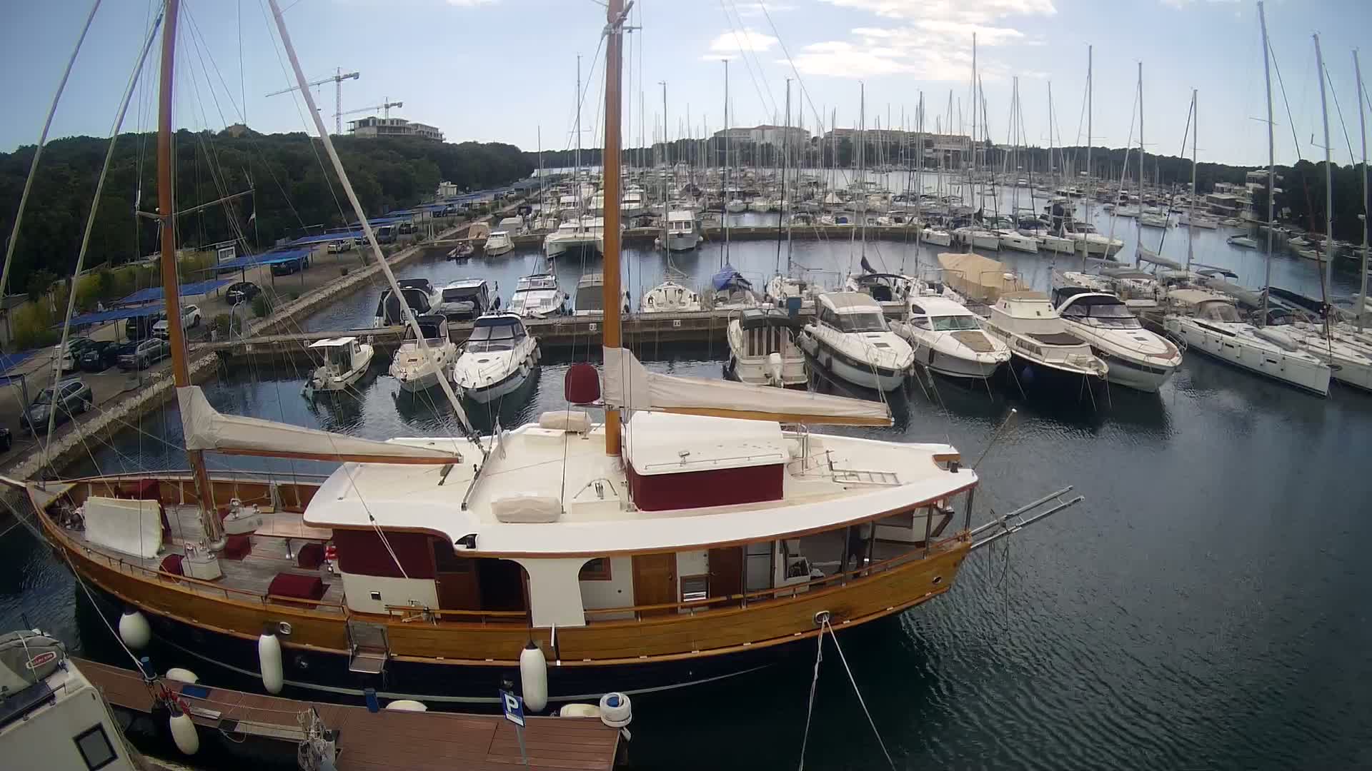 A large wooden sailboat is docked in a marina filled with numerous other sailboats and motorboats on a mostly sunny day.
