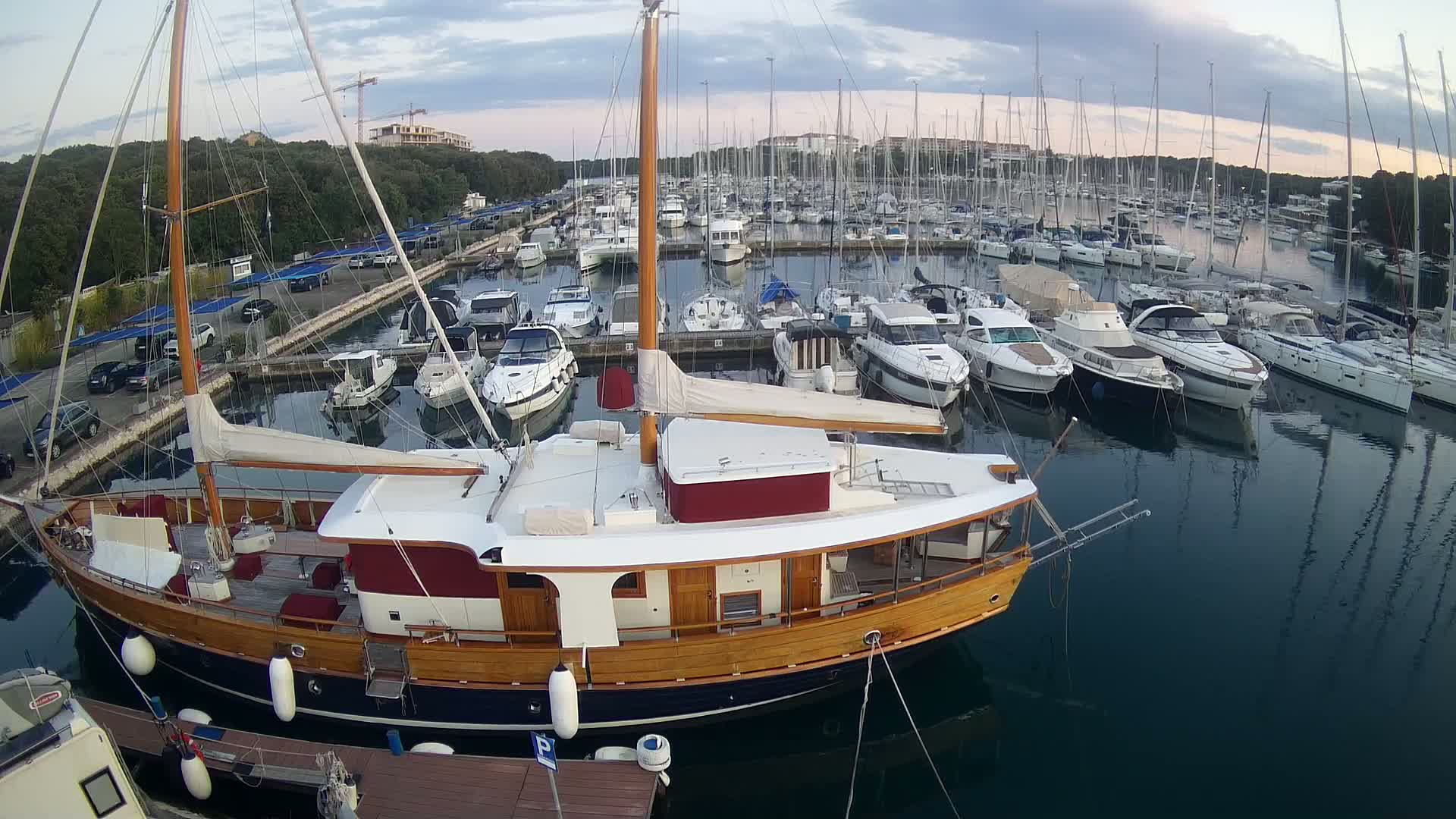 A large wooden sailboat is docked in a marina filled with numerous other sailboats and motorboats under a partly cloudy sky.
