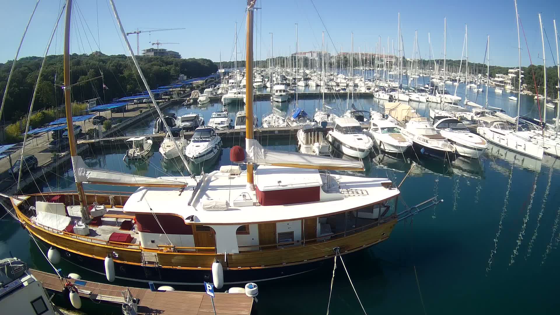 A large wooden sailboat is docked in a marina filled with numerous other sailboats and motorboats on a sunny day.