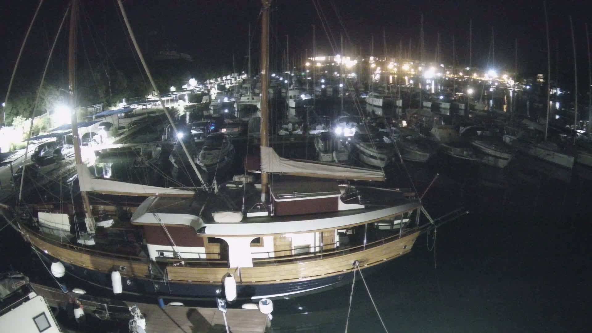 A large wooden sailboat is docked at night in a marina filled with numerous other boats.