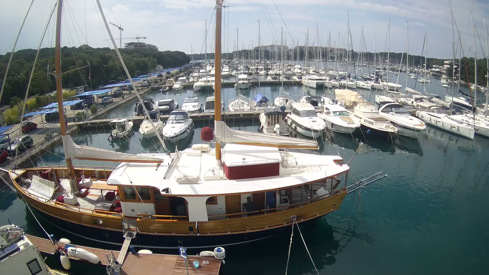 A large wooden sailboat is docked in a marina filled with numerous other boats on a sunny day.