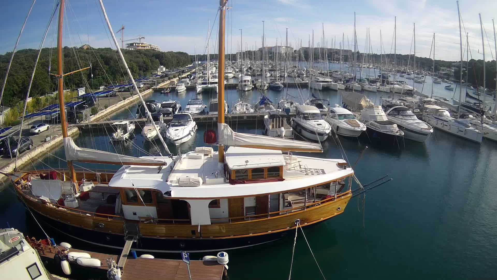 A large wooden sailboat is docked in a marina filled with numerous other boats on a sunny day.