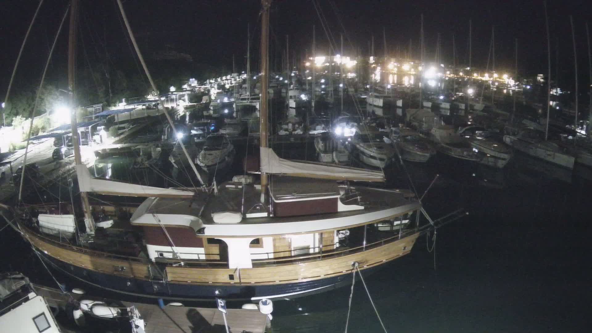 A large wooden sailboat is docked at night in a marina filled with many other boats under dark skies.
