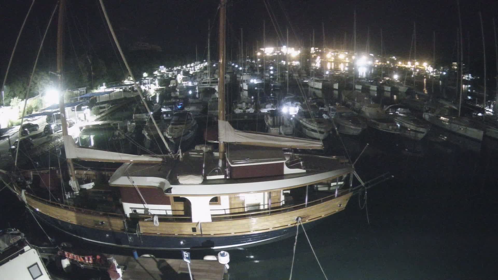 A large wooden sailboat is moored in a marina at night, surrounded by numerous other boats under dark, clear skies.