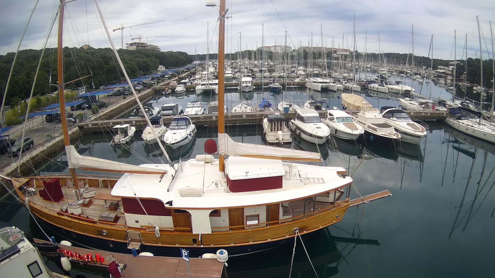 A large wooden sailboat is moored in a marina filled with numerous other sailboats and motorboats under a cloudy sky.