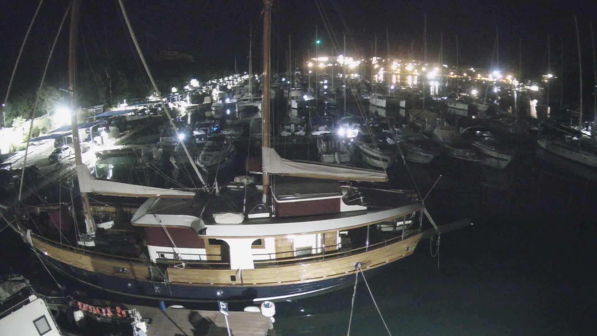 A large wooden sailboat is docked at night in a marina filled with many other boats under a clear, dark sky.