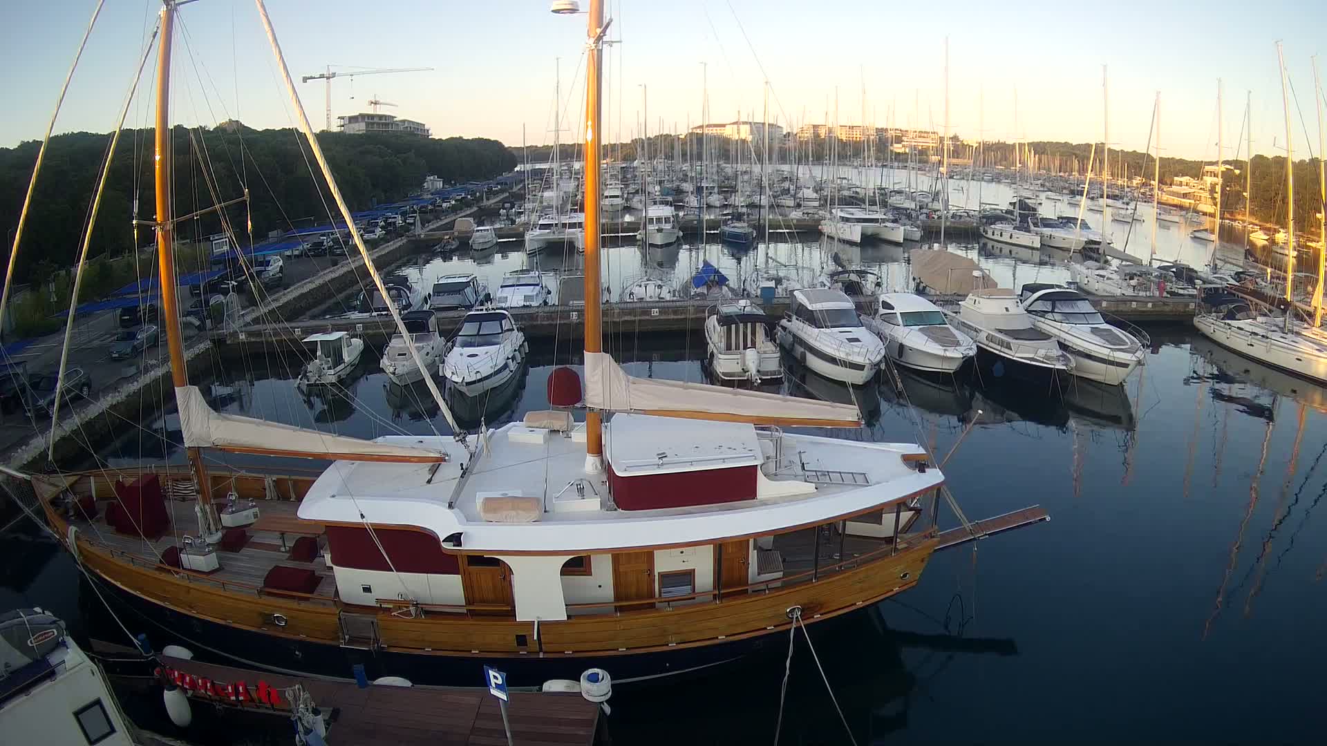 A large wooden sailboat is docked in a marina filled with numerous other boats on a calm, clear morning.