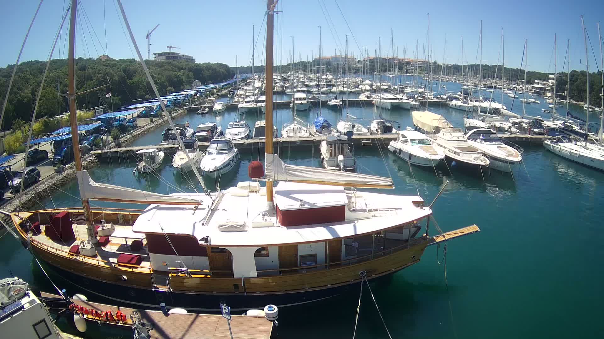 A large wooden sailboat is docked in a marina filled with numerous other boats on a sunny day.