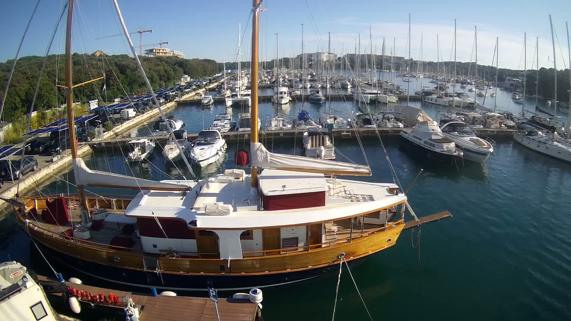 A large wooden sailboat is docked in a marina filled with many other boats on a sunny day.