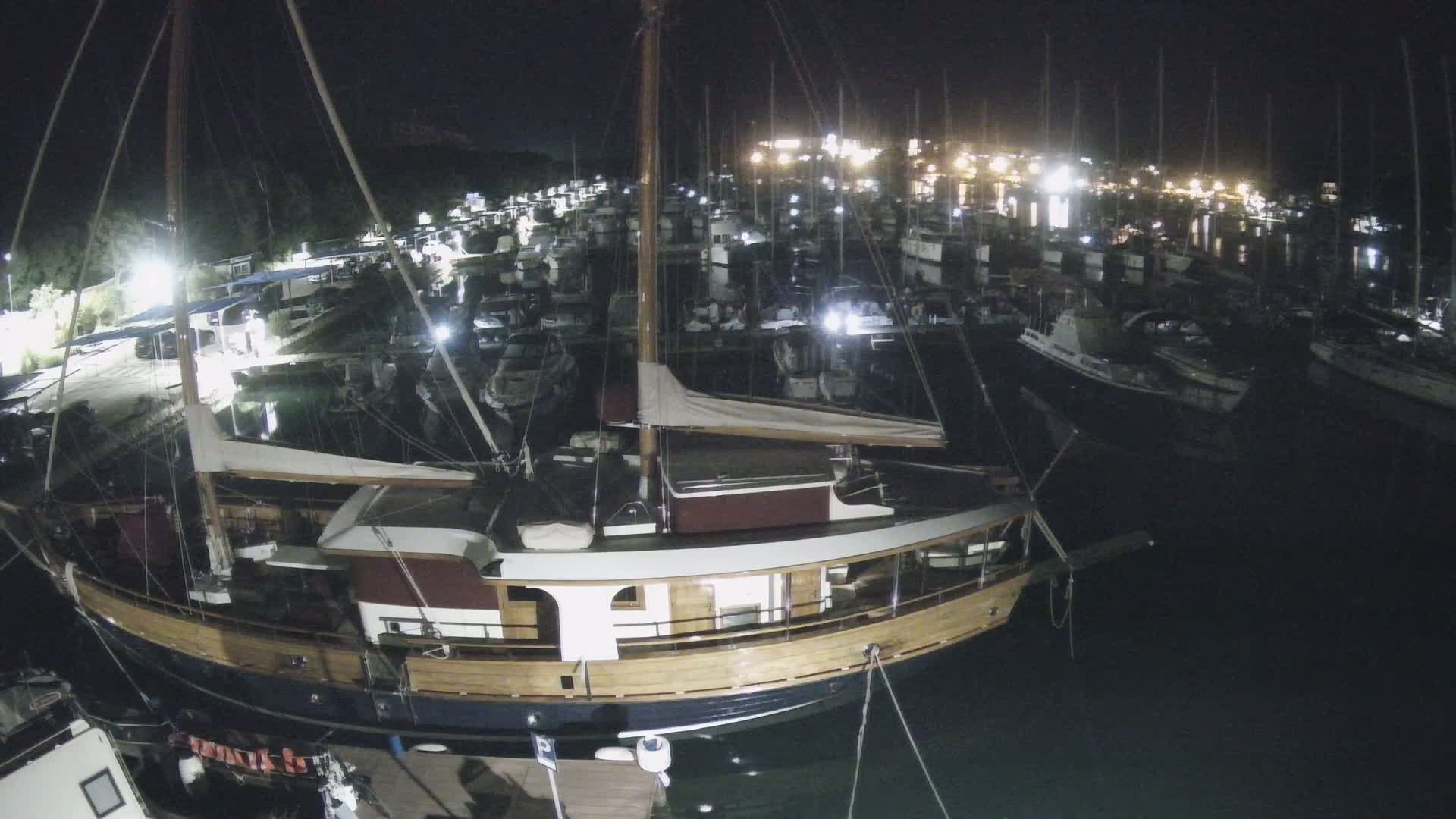 A large wooden sailboat is docked at night in a marina filled with many other boats under dark, clear skies.