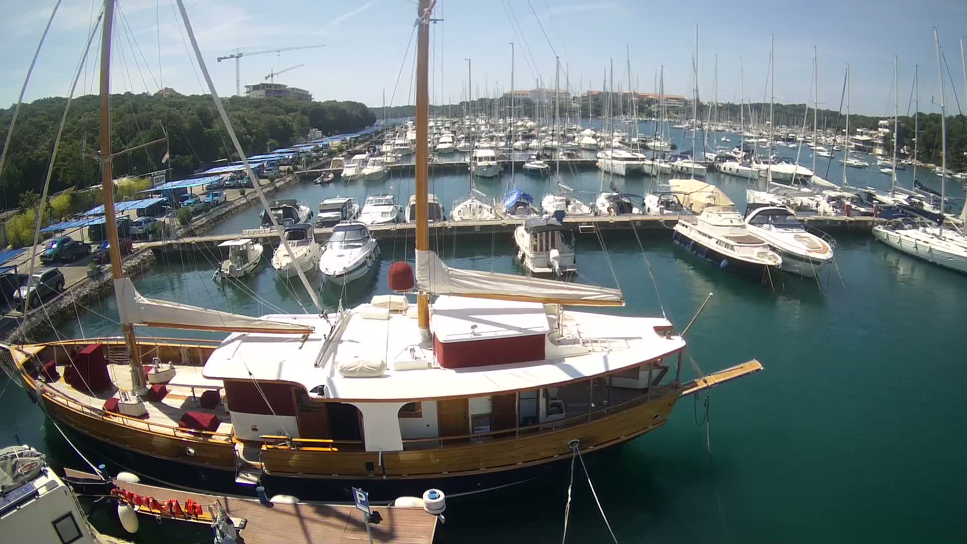 A wooden sailboat is moored in a marina filled with numerous other sailboats and motorboats on a sunny day.