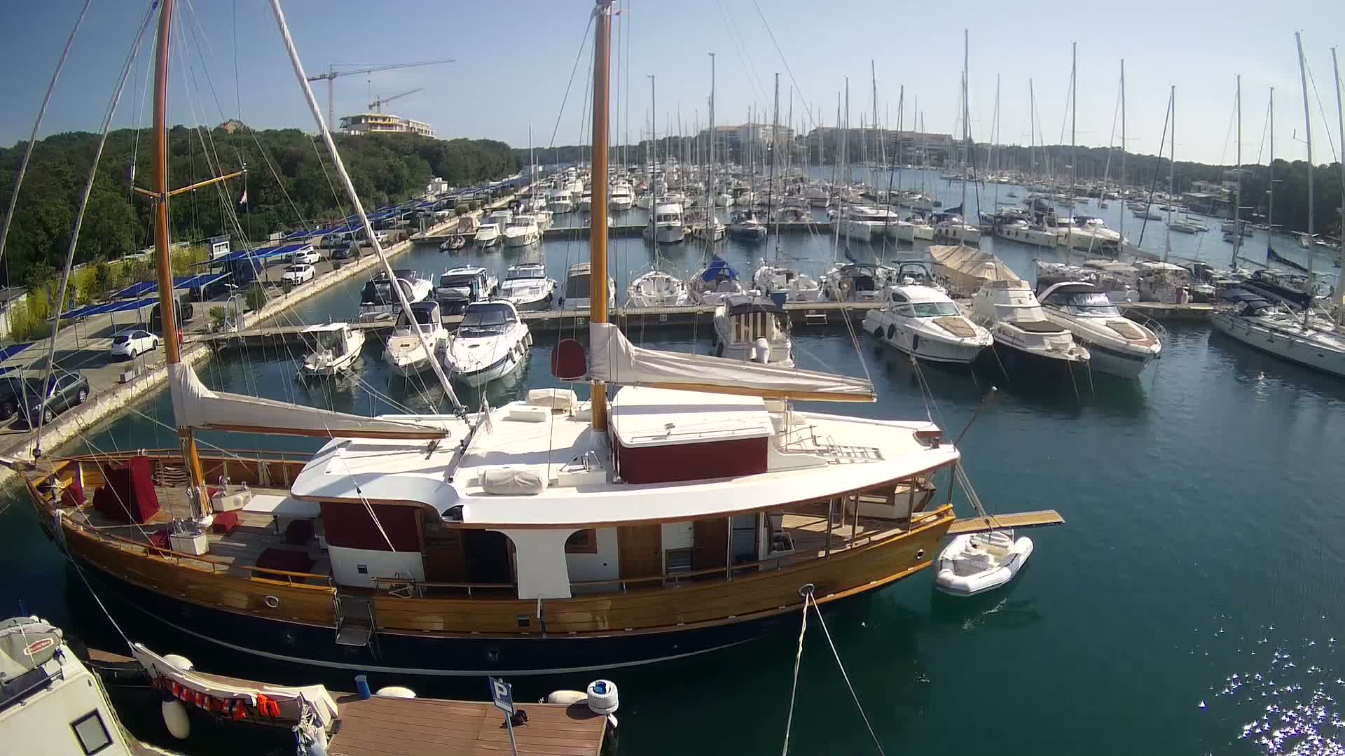 A large wooden sailboat is docked in a marina filled with numerous other boats on a sunny day.