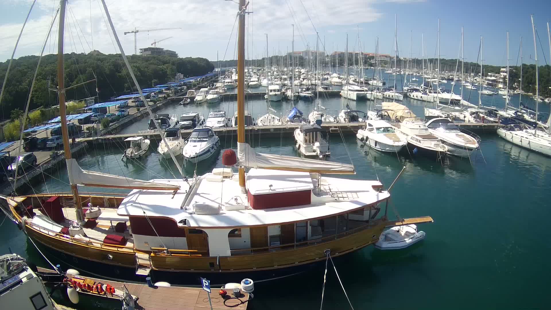 A large wooden sailboat is docked in a marina filled with numerous other boats on a sunny day.