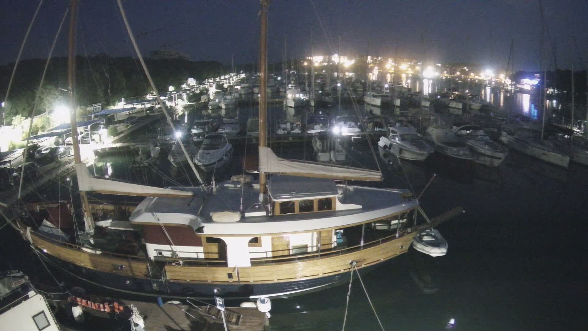 A large wooden boat is moored in a marina at night, surrounded by numerous other boats and illuminated by lights on shore.